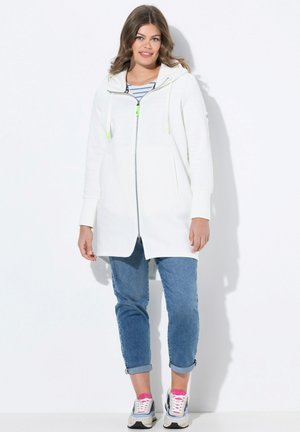 Weiße Zip-Jacke mit Kapuze, ausgestattet mit neongrünen Akzenten, Seitentaschen und einem lockeren Schnitt. Kombiniert mit aufgerollten Jeans und Sneakers.