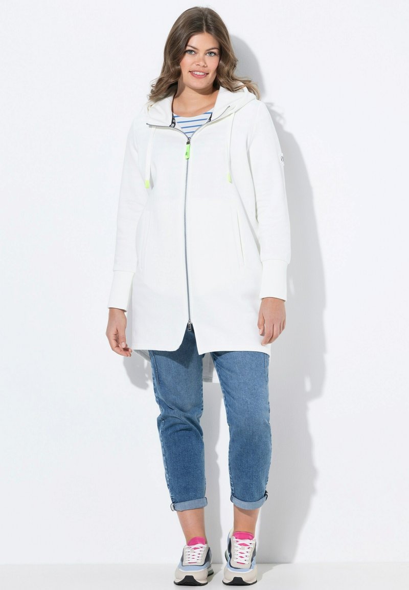 Bílá bunda na zip s kapucí, obsahující neonově zelené akcenty, postranní kapsy a uvolněný střih. V kombinaci s ohrnutými denimovými kalhotami a teniskami.