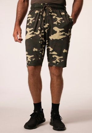 Mand iført grønne og beige camouflageshorts, sorte sokker og sorte sportssko, står med den ene hånd i lommen.