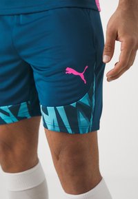 Blaue sportliche Shorts mit einem pinken Puma-Logo, versehen mit einem geometrischen türkisfarbenen Muster am Saum und aus weichem, atmungsaktivem Stoff.