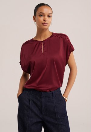 Femme portant un chemisier en satin bordeaux à manches courtes avec un col en trou de serrure et un pantalon sombre taille haute, debout les mains dans les poches.