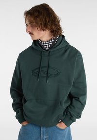 Felpa con cappuccio verde scuro con logo testurizzato, tasca a marsupio e cappuccio con coulisse. Indossata sopra una sciarpa a scacchi bianca e nera.