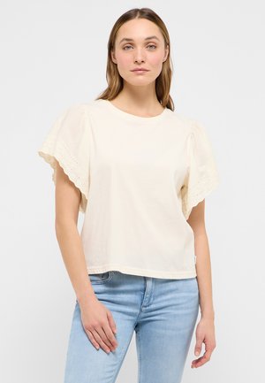 Frau trägt eine cremefarbene Bluse mit weit geschnittenen, gewellten Spitzensäumen an den Ärmeln und hellblaue Jeans, steht vor einem schlichten weißen Hintergrund.