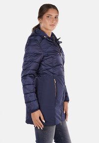 Giacca imbottita navy con zip frontale, design aderente e cappuccio. Presenta tasche laterali con dettagli in hardware dorato e una consistenza liscia.