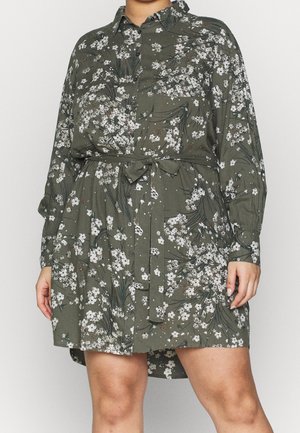 Personne portant une robe fleurie olive à manches longues, boutonnée, avec une ceinture à la taille et un imprimé de fleurs blanches, montrée du cou aux cuisses.