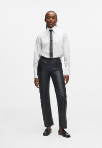 Λευκό πουκάμισο με κουμπιά και αντιθετικό μαύρο γραβάτα, συνδυασμένο με κομψά μαύρα παντελόνια από γυαλιστερό ύφασμα και μαύρες λοφάδες.