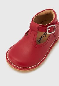 Κόκκινο παιδικό δερμάτινο παπούτσι με λεπτομέρεια κυματιστής άκρης, πλευρικό λουράκι με αγκράφα, μπεζ ραφές και ταμπά σόλα σε λευκό φόντο.