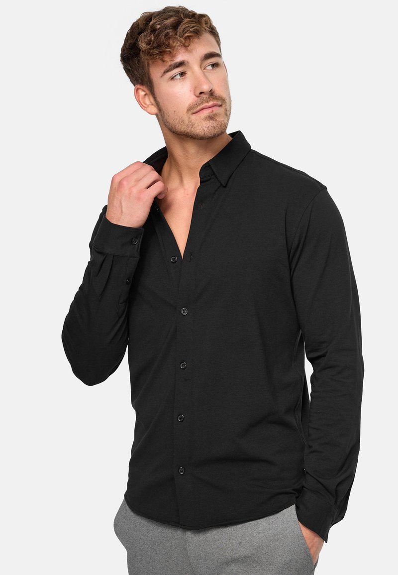 Camisa negra de manga larga con botones y cuello clásico, con una textura suave y un diseño ajustado, presentada con el cuello desabrochado.