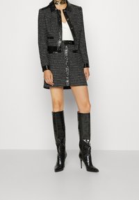 Zwarte tweed blazer met glanzende zwarte lakkraag en knopen, gecombineerd met een bijpassende tweed rok. Knielange zwarte laarzen van lakleer met krokodillenpatroon.