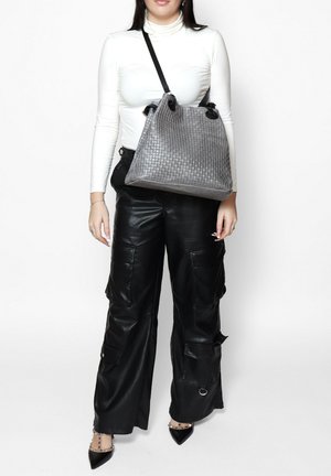 Grijze geweven leren emmer tas met zwarte handvatten, met een gestructureerd oppervlak en ronde sluitingen. Gestyled met een witte coltrui en zwarte leren broek.
