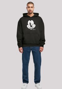 Felpa nera con una grande grafica di Topolino bianco sul fronte, dotata di tasca a marsupio e polsini a coste, abbinata a jeans blu e sneakers bianche.