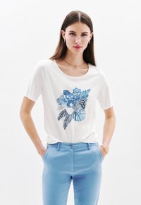Witte t-shirt met korte mouwen, voorzien van een blauw bloem- en bladmotief, gecombineerd met lichtblauwe broek. Het heeft een ronde hals en een zachte textuur.