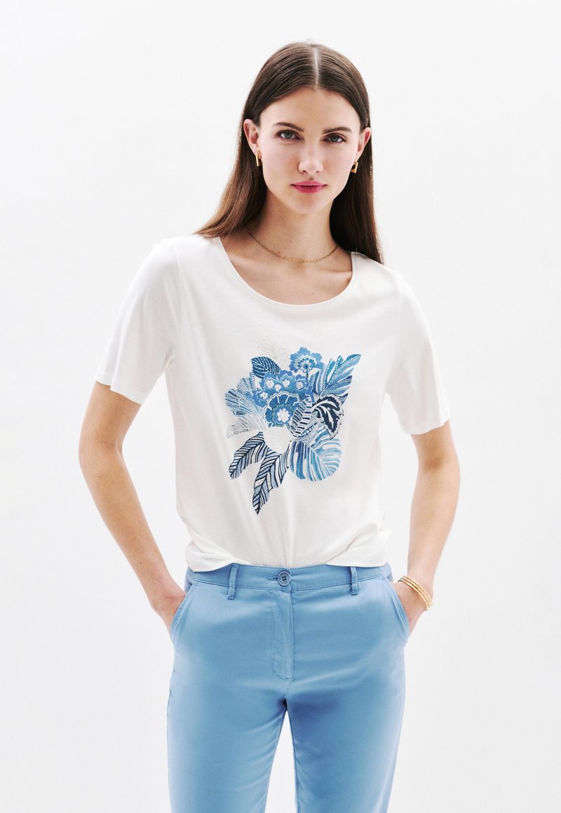 Witte t-shirt met korte mouwen, voorzien van een blauw bloem- en bladmotief, gecombineerd met lichtblauwe broek. Het heeft een ronde hals en een zachte textuur.