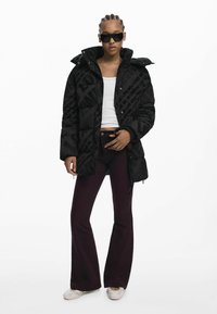 Veste doudoune noire avec des accents brillants et un motif textuel répété "LOVE ME", associée à un pantalon bordeaux évasé et des chaussures à enfiler claires.