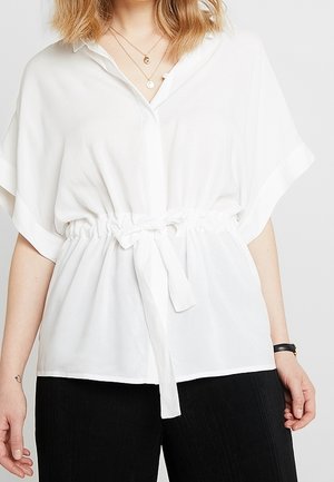 Woman wearing a loose white short-sleeve blouse with a cinched waist tie, layered gold necklaces, black pants, and a black wristwatch.