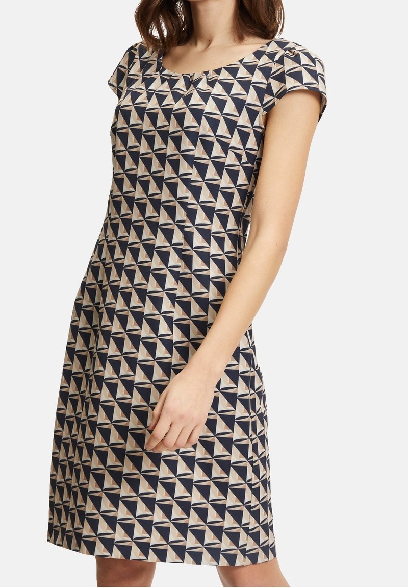 Femme portant une robe courte à manches courtes avec un motif géométrique de triangles bleu marine, beige et blanc sur un fond clair.