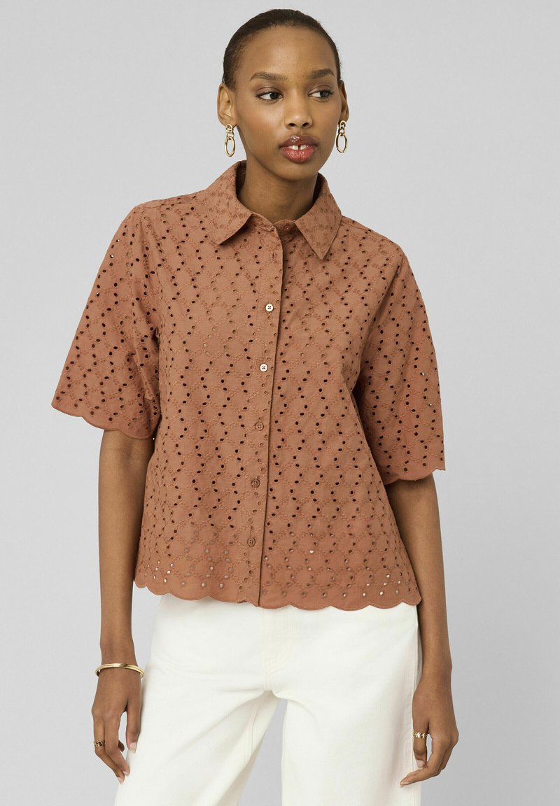 Femme portant un chemisier marron à œillets à manches courtes avec des bords festonnés et un pantalon blanc, accessoirisée avec des boucles d'oreilles créoles en or et des bagues.