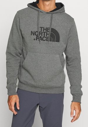 Mężczyzna w szarej bluzie z kapturem The North Face z przednią kieszenią i sznurkami, w zestawieniu z ciemnoszarymi spodniami, na jednolitym tle.