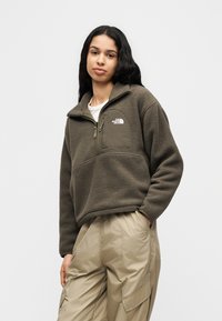 Νέα γυναίκα με μακριά σκούρα μαλλιά, που φορά καφέ φλις πουλόβερ με τσέπη στο στήθος και ανοιχτό μπεζ παντελόνι cargo, στέκεται με το ένα χέρι στην τσέπη.