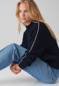 Pull en maille bleu marine avec un col haut et des rayures blanches sur les côtés, associé à un jean en denim bleu clair. Texture douce et coupe décontractée.