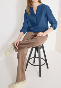 Femme portant une chemise bleu à manches longues, un pantalon marron et des baskets blanches, assise en tailleur sur un tabouret noir, sur fond blanc.
