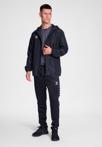 Chaqueta negra ligera con capucha, combinada con pantalones deportivos negros que presentan rayas en forma de chevrón. El material parece suave con puños elásticos.