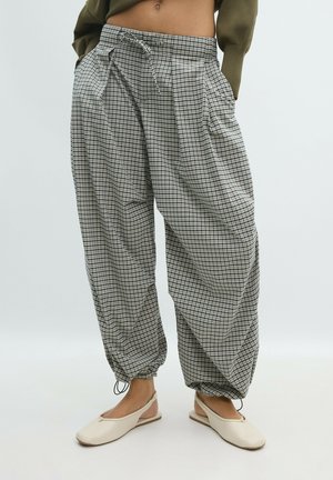 Personne portant un pantalon ample à taille haute à carreaux avec cordon de serrage et poignets froncés, associée à des chaussures plates beiges à bride arrière.