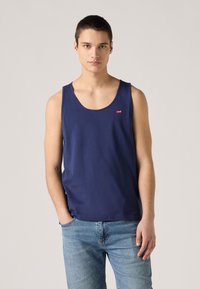Tumesinine puuvillane ilma varrukatops, ümar kaelus ja väike punane logo pael ees, koos helesiniste denim teksadega.
