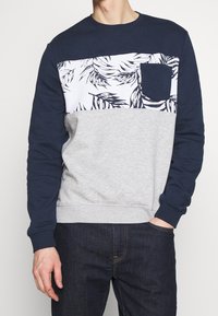 Joven vestido con un sudadero azul marino y gris que tiene un panel en el pecho con un patrón floral blanco y un bolsillo, combinado con jeans oscuros.