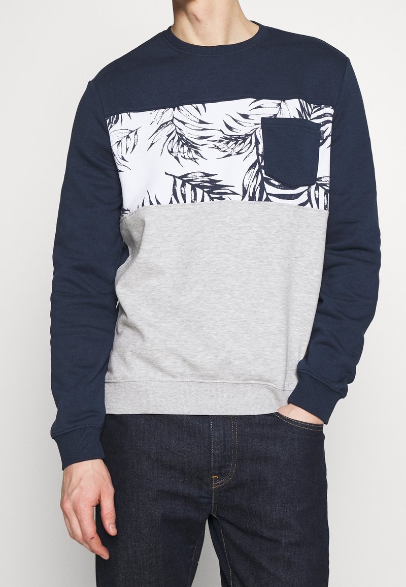 Joven vestido con un sudadero azul marino y gris que tiene un panel en el pecho con un patrón floral blanco y un bolsillo, combinado con jeans oscuros.