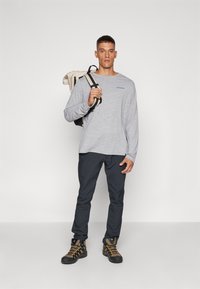 Siva majica dugih rukava s plavim logom, tamne hlače slim fit kroja i planinarske čizme s žutim detaljima. Model nosi bež i crni ruksak.