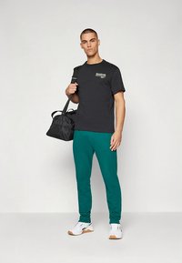 Schwarzes T-Shirt mit Logo, grüne Jogginghosen, hält eine schwarze Sporttasche. Modell trägt weiße Turnschuhe; neutraler Hintergrund.