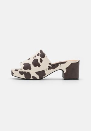 Plateau-Mules mit Kuhmuster in Schwarz und Beige, mit glatter Textur, offenem Zehenbereich und klobigem Absatz für Stabilität.