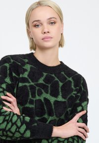 Schwarzer und grüner Pullover mit einem strukturierten Muster, das unregelmäßige Formen ähnelt, mit Rundhalsausschnitt und langen Ärmeln. Weiches, flauschiges Material.