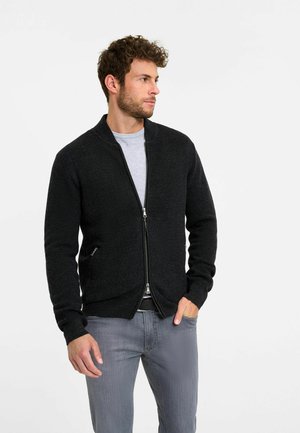 Schwarze Strickjacke mit Reißverschluss, gerippter Textur und rundem Kragen. Kombiniert mit einem grauen T-Shirt und grauen Slim-Fit-Jeans.