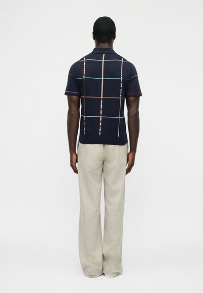 Homme debout, de dos, portant un polo à manches courtes bleu marine avec des lignes de grille multicolores et un pantalon beige à jambes larges.