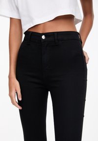 Jeans negros de talle alto de denim, con cierre frontal de botón, trabillas para el cinturón y una textura suave y elástica.