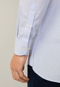Camicia a quadretti azzurro chiaro con polsini abbottonati, caratterizzata da una texture liscia e un design a orlo curvo. I bottoni sono bianchi e arrotondati.