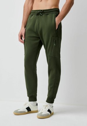 Homme portant un pantalon de jogging vert olive avec poches latérales zippées et des baskets blanches à rayures noires, debout avec une main dans la poche.