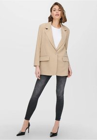 Beżowa dopasowana marynarka z wyłogami, z dwoma przednimi kieszeniami, zamykana na pojedynczy guzik, noszona na białej koszuli, zestawiona z ciemnymi wąskimi dżinsami.