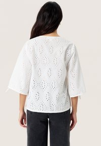 Hvid øjelet bluse med diamantudskæringer og klokkeærmer, med en afslappet pasform og en rund halsudskæring, vist mod en neutral baggrund.