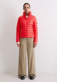 Czerwona podszyta kurtka z wysokim kołnierzem, całkowitym zamkiem błyskawicznym i poziomym przeszyciem. Połączona z beżowymi szerokimi spodniami i czarnymi butami.