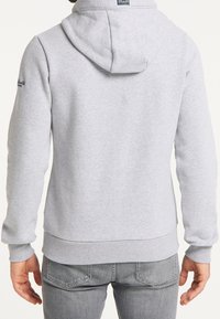 Homme portant un sweat à capuche gris clair et un jean gris, de dos, les mains détendues le long du corps, sur fond blanc.