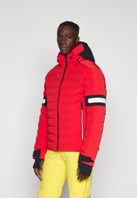 Rote Pufferjacke mit Kapuze, ausgestattet mit schwarzen Akzenten, weißen Streifen an den Ärmeln, einem Logo auf der Brust und Reißverschlusstaschen. Strukturierte Oberfläche.