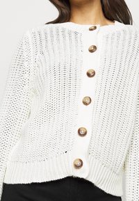 Cardigan tricoté blanc avec un motif texturé, encolure ronde et boutons marron à l'avant. Ourlet et poignets côtelés.