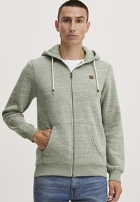 Haut à capuche zippé vert clair en tissu texturé, avec une poche avant, un cordon de serrage sur la capuche et un petit patch logo marron sur la poitrine.