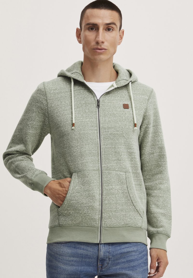 Haut à capuche zippé vert clair en tissu texturé, avec une poche avant, un cordon de serrage sur la capuche et un petit patch logo marron sur la poitrine.