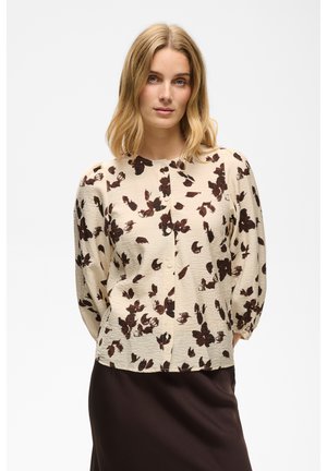 Kvinde med skulderlangt blondt hår iført en beige bluse med mørkebrun blomsterprint og en mørkebrun nederdel, stående med hænderne bag ryggen.