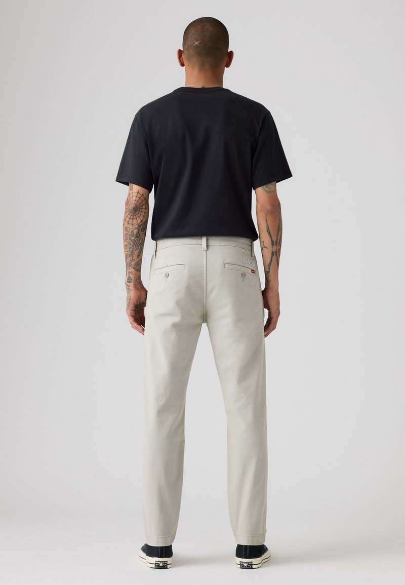 Homme aux bras tatoués portant un t-shirt noir, un pantalon beige clair avec poches arrière et des baskets noires, debout, dos tourné, sur un fond uni.