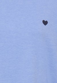Pull tricoté bleu clair avec un petit emblème en forme de cœur noir sur le côté gauche. Texture douce et design simple avec tissu côtelé.
